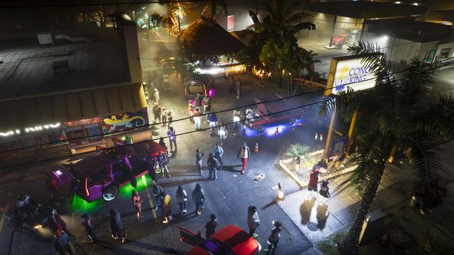 A crowd gathers at night around cars with glowing underlights in a lot beside a Conch King sign