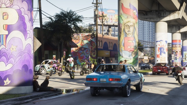 A blue sedan on big chrome wheels rolls under a mural-covered overpass in Vice City as riders on dirt bikes and ATVs pop wheelies at the intersection ahead.