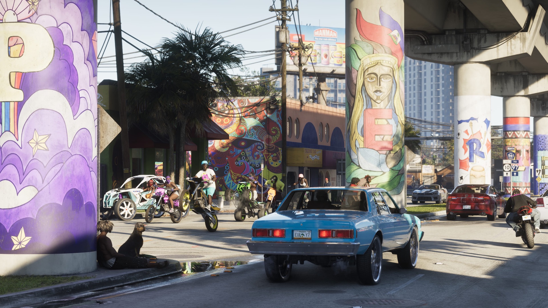 A blue sedan on big chrome wheels rolls under a mural-covered overpass in Vice City as riders on dirt bikes and ATVs pop wheelies at the intersection ahead.